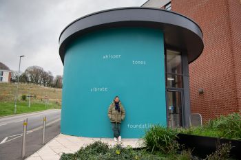 Helen Cammock stands in front of the artwork on an external wall at top of СƵ Student Centre. The text is painted in white and set against a bright teal background, and reads: whisper  tones  vibrate  foundations